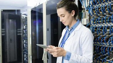 Frau im Laborkittel arbeitet in einem Serverraum, umgeben von technischen Geräten und Kabeln, fokussiert auf ihre Arbeit.