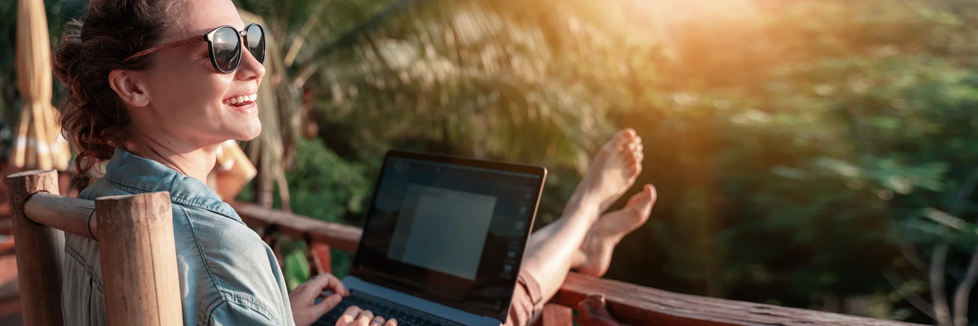 Eine lachende Frau sitzt auf einem Balkon mit Blick auf einen Wald udn arbeitet an einem Laptop.