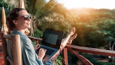 Eine lachende Frau sitzt auf einem Balkon mit Blick auf einen Wald udn arbeitet an einem Laptop.