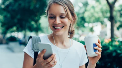 Eine Frau geht durch einen Park und schaut lächelnd auf ihr Handy in der rechten Hand. In der linken Hand hält sie einen Kaffee-Becher
