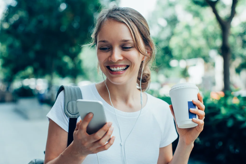 Eine Frau geht durch einen Park und schaut lächelnd auf ihr Handy in der rechten Hand. In der linken Hand hält sie einen Kaffee-Becher